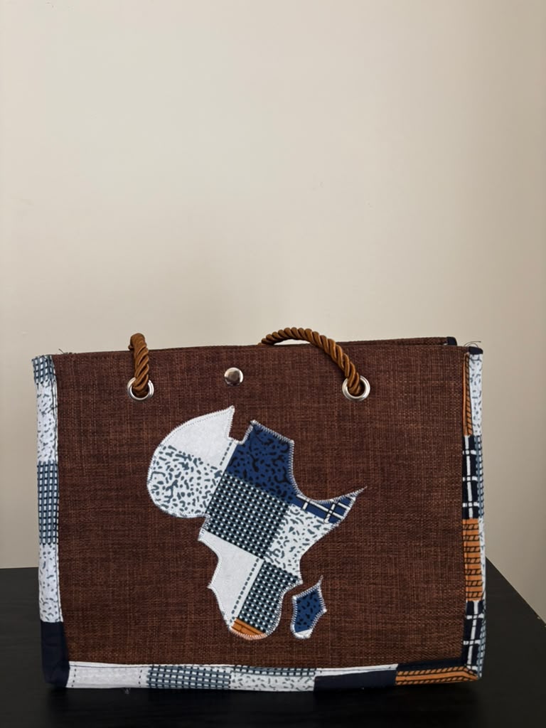A brown woven tote bag featuring a patchwork-style map of Africa on the front in white, blue, and navy tones, with rope handles and patterned fabric trim, shown against a plain beige wall.